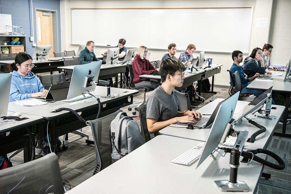 Students sit at desks in a classroom with laptops and computer monitors.