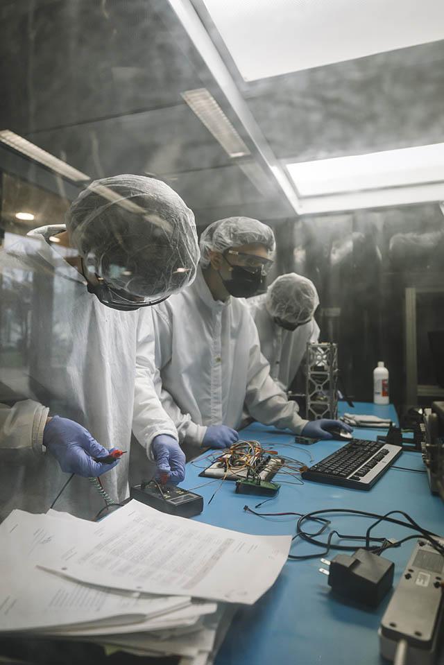 Three students wearing protective lab gear work at a table with various pieces of equipment and a computer.